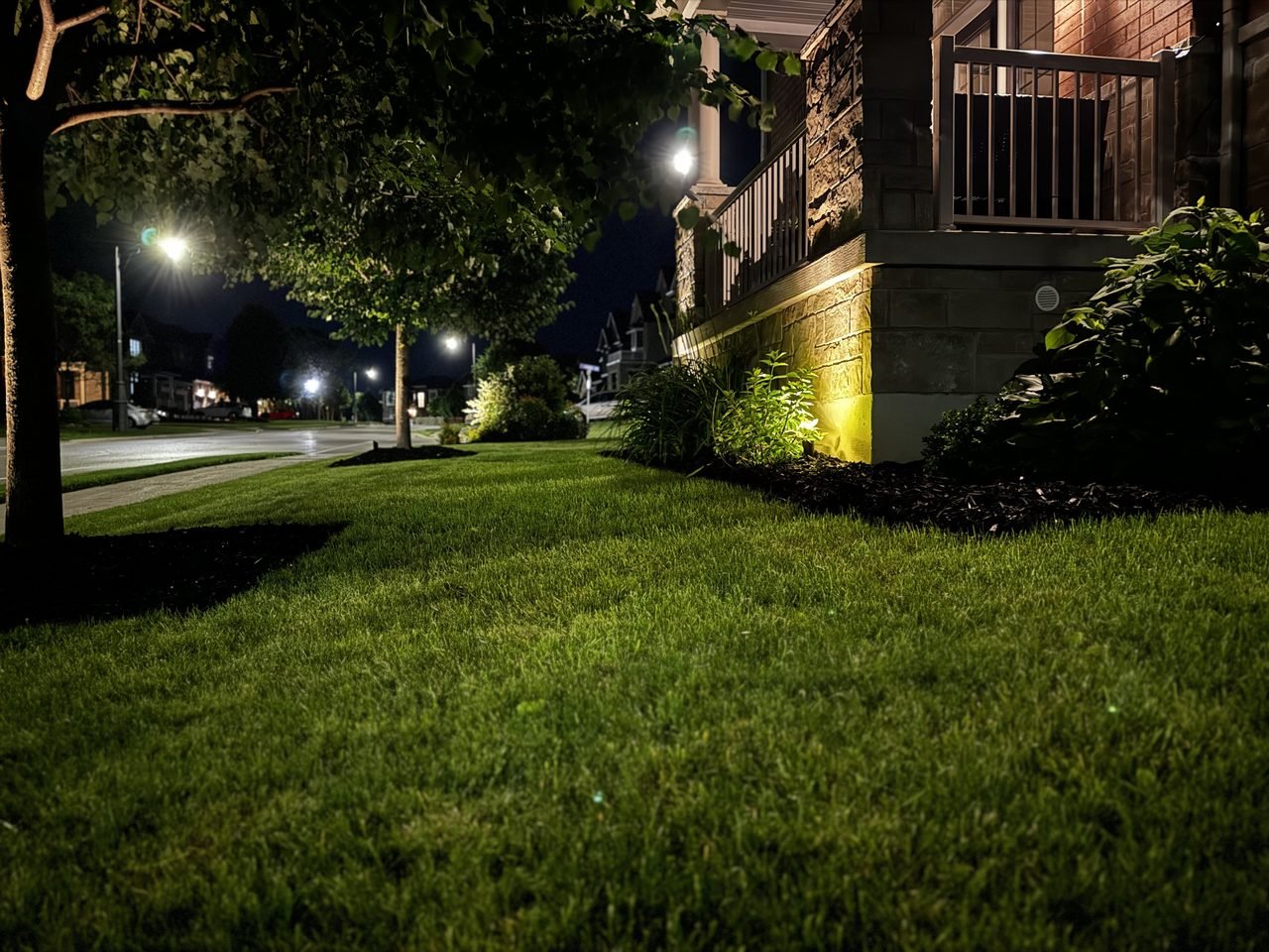 A GTA home at night with warm uplighting washing up the stone facade and lit garden beds