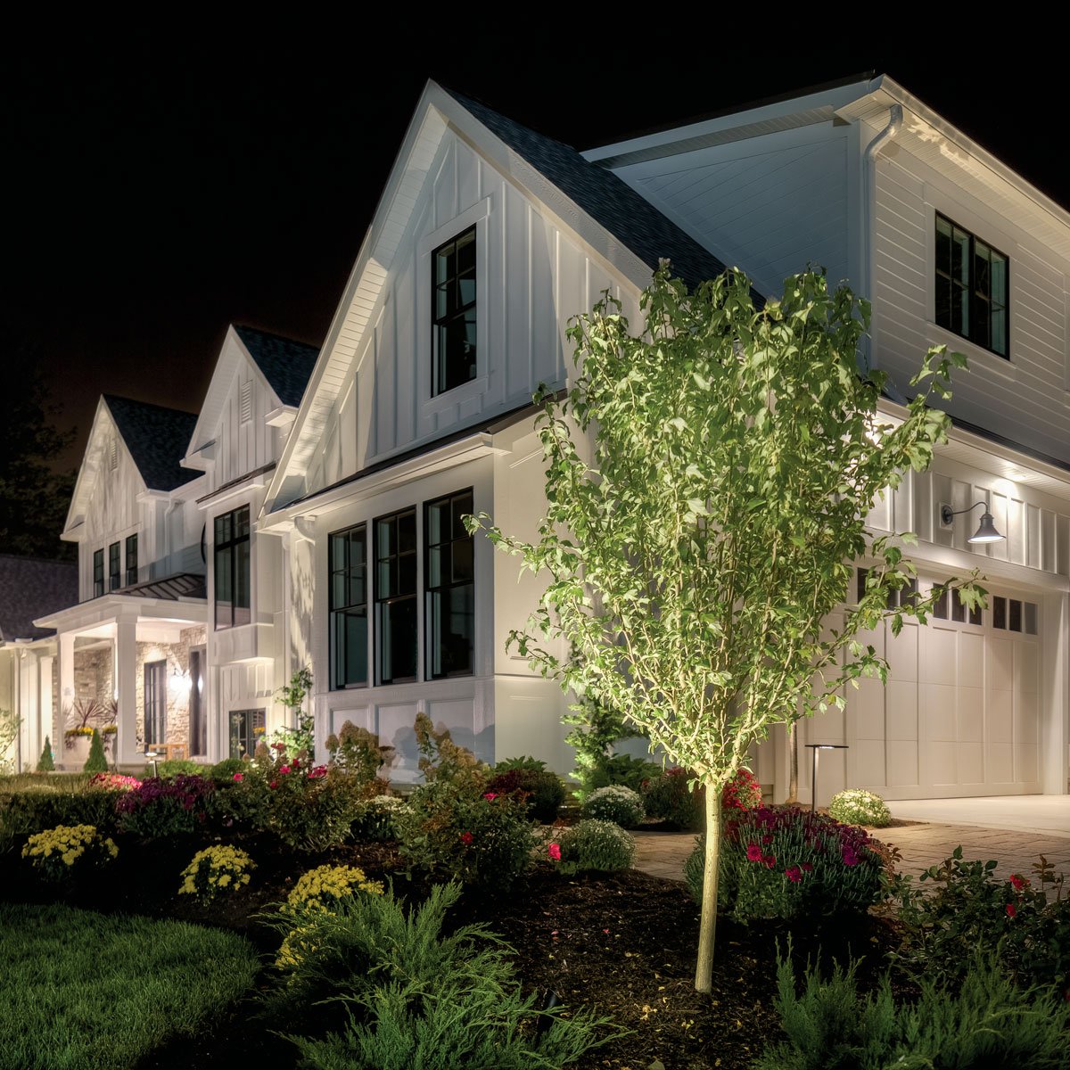 A white farmhouse-style home at night with driveway path lights and an uplit tree