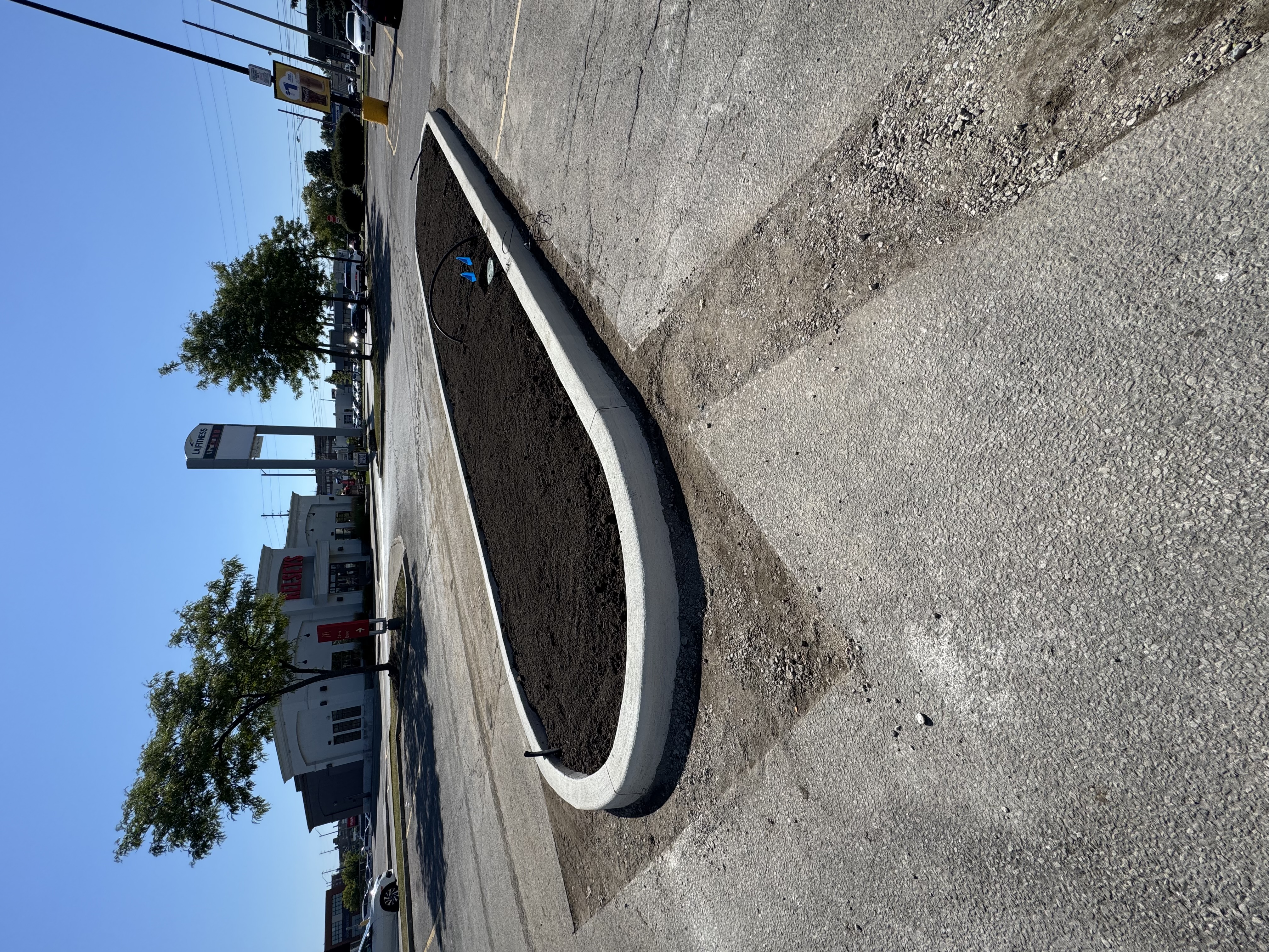Commercial parking-lot planter island prepped for planting — concrete curb, fresh soil, drip line laid with marker flags, mini excavator on site