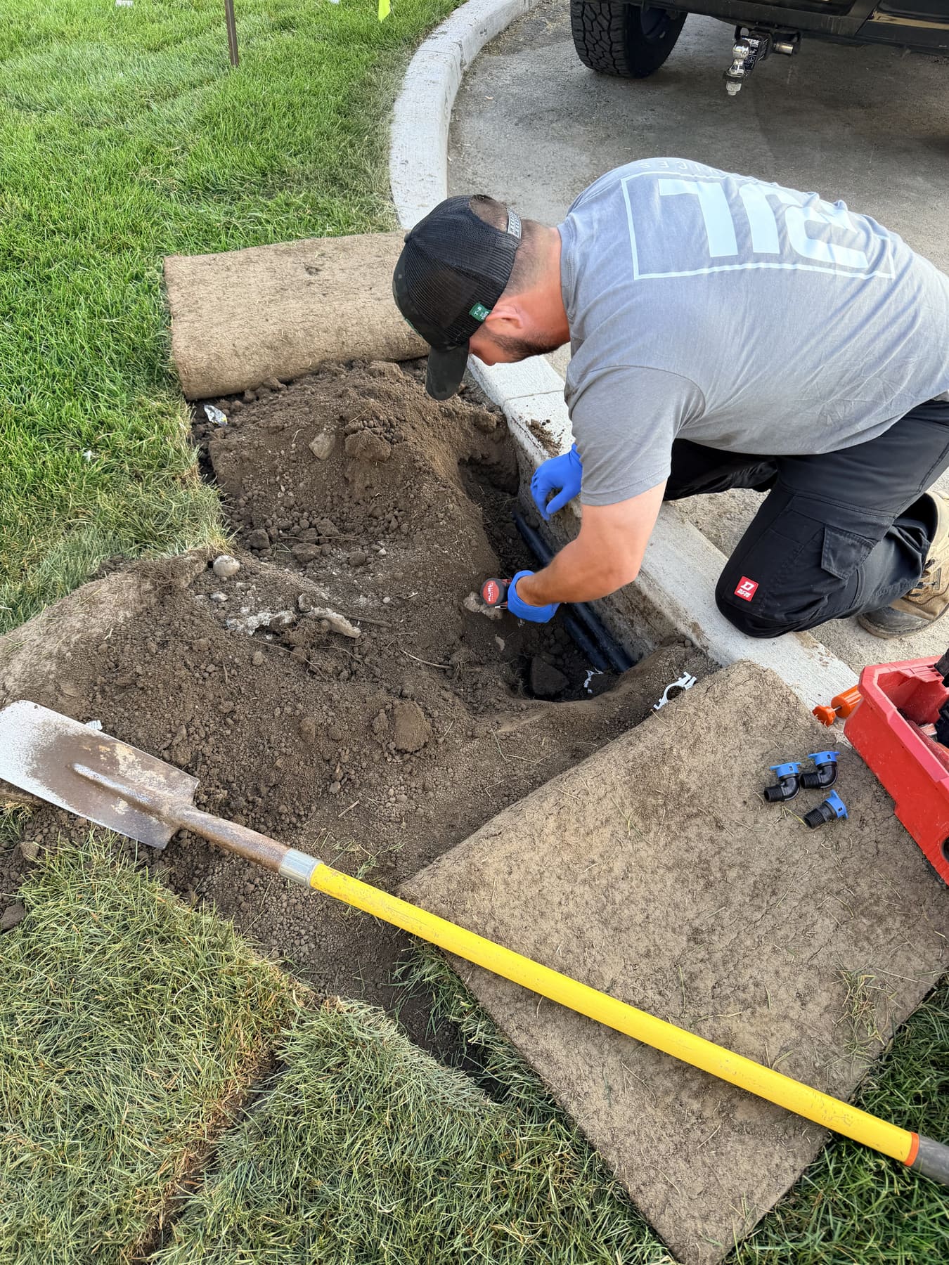Overhead of a PJL Land Services technician repairing an irrigation mainline at a curbside trench, sod cut back and tools laid out
