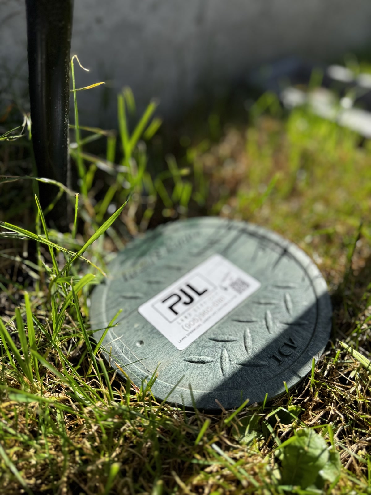 Close-up of a PJL Land Services-branded valve box cover with company logo, phone number, and service QR code, installed in a residential lawn.